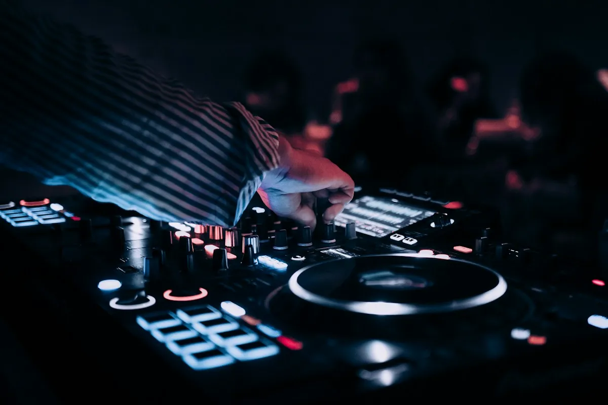 A DJ adjusting a lit-up controller with guests dancing in the background at an evening work party